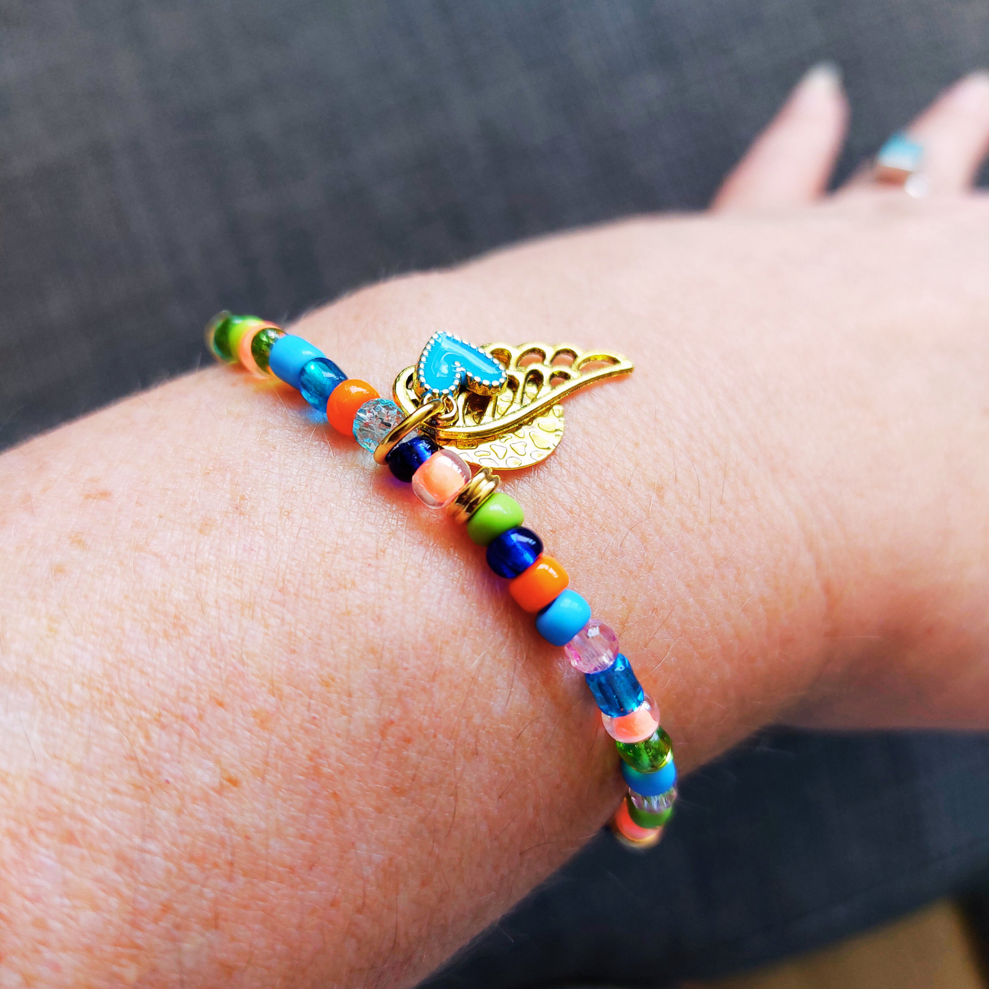Armband elastisch in met rocailles en facetkralen in frisse groene, blauwe tinten met een vleugje roze en oranje. Bedels vleugel en munt in goudkleur. Foto is sfeerbeeld van armband om pols.